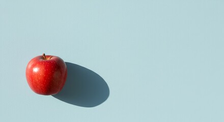 A vibrant red apple casts a stark shadow against a pastel blue backdrop, presenting a clean, minimalist aesthetic.