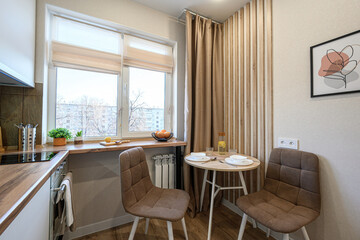 Cozy kitchen corner with a round table set for two, lit by a window. Neutral tones with wood accents create a warm, inviting atmosphere