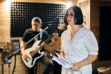 Young woman with black bob haircut sings at microphone, wearing stylish white dress, holding papers. Male guitarist with cap focused on instrument. Warm lighting, wooden room, soundproof walls