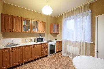 cozy kitchen featuring wooden cabinets, a white table, and a window with sheer curtains. Neutral tones dominate, with warm lighting