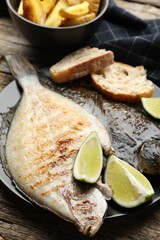 Tasty roasted flounder fish served with potatoes, bread and lime slices on wooden table, closeup. Homemade seafood dish