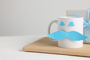 Happy Father's Day. Cup with paper face and gift box on white wooden table, closeup. Space for text