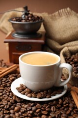 Cup of hot coffee, beans, grinder and cinnamon on brown background, closeup