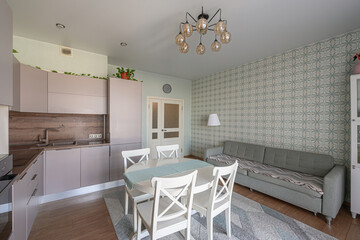 cozy apartment scene with a blend of kitchen and living spaces. It features mauve cabinets, a white table with chairs, a grey sofa, and patterned wallpaper