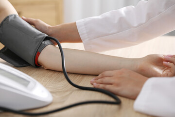 Doctor checking woman's heart rate and blood pressure via sphygmomanometer at wooden table indoors, closeup
