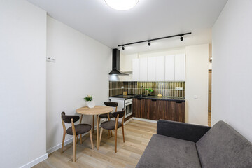 bright studio apartment with a kitchenette, dining table, and sofa. The kitchen features white cabinets and dark wood lower cabinets