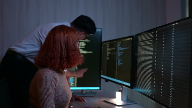 Professional programmers analyzing code on multiple monitors in dark office. Man and woman developers collaborating on cybersecurity project late at night in modern technology workplace.