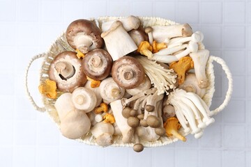Different raw mushrooms in wicker basket on white tiled table, top view