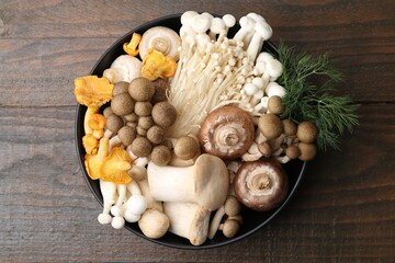Different raw mushrooms and dill in bowl on wooden table, top view