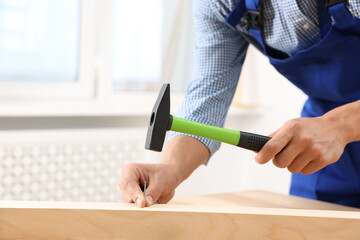 Professional repairman hammering nail into wooden plank indoors, closeup. Space for text