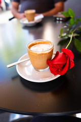 Glass of cortado coffee with red rose on saucer and spoon on a black table