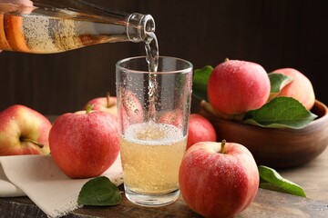 Pouring delicious apple cider from bottle into glass at wooden table against dark background, closeup