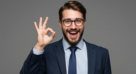 Smiling businessman with glasses suit tie makes OK hand gesture against a plain gray background
