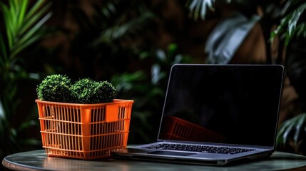 Orange shopping basket with plants sits beside a laptop on a dark table, lush greenery in background.