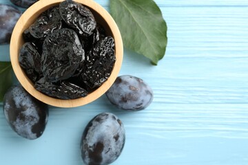Dried prunes in bowl, leaves and fresh plums on light blue wooden table, flat lay. Space for text