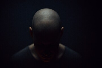 Bald man with head tilted downward, dramatic low-key photography style, dark background, concept of solitude and introspection