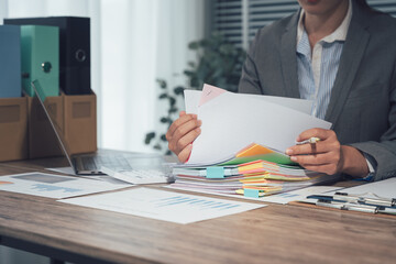 Businessman working through pile of documents to find unfinished documents, information on pile of documents on desk and checking financial documents amidst busy workload.