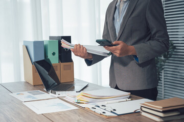 Business Document and Device: A professional individual, smartly attired in a business suit, meticulously reviews documents and utilizes a digital device on a well-organized desk.