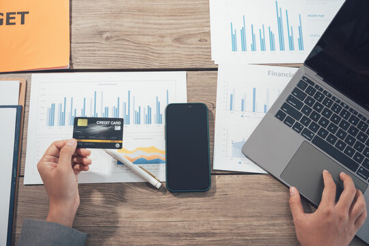 Financial Planning in Progress: A close-up shot captures the meticulous process of financial planning, with a hand holding a credit card poised above a smartphone and laptop. surrounded by graphs.