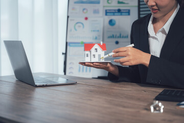 Property Presentation: A professional presents a miniature house, possibly showcasing real estate options or design concepts, complemented by a laptop on a wooden desk.