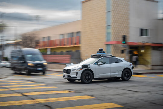LOS ANGELES, CA - March 11, 2025: Google's Alphabet Waymo driverless self driving taxi cab rushing through city traffic, crossing street intersection with Amazon Prime delivery van in the background.