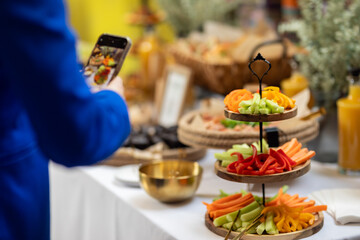 Close-up of fresh vegetable snacks arranged on a tiered wooden server, with a guest capturing the moment at a catered event