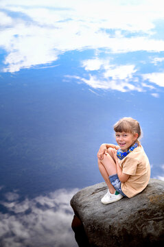 Fototapeta A little cute girl sitting on a rock near the lake. Sky reflection in the water.