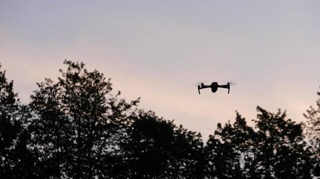 The silhouette of a drone hovering in the air against the backdrop of dark trees
