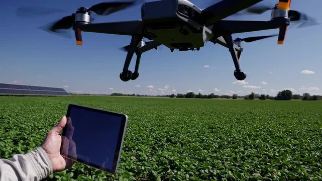 Aerial view of a drone flying over a lush green field, with a person holding a tablet, capturing video footage of agricultural technology.