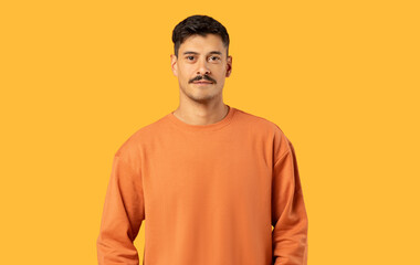 A young man in an orange sweater stands calmly with a neutral facial expression against a solid yellow background © Prostock-studio