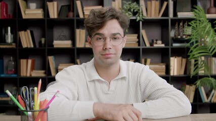 A boy student sitting on a bench in a classroom looks directly at the camera,smiles and demonstratively raises his hand up as a sign that he wants to answer, to give his opinion.Good manners at school