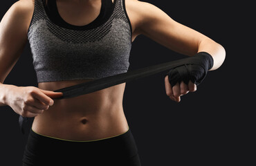 Athletic female boxer wrapping hands with boxing tape before fight. Female fitness model with muscular body at black studio background, low key. Bodybuilding concept, copy space