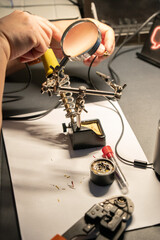 Close-up of hands soldering a cable with a magnifying glass and tools on a workbench