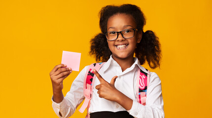 Cute Black Elementary Student Girl Pointing Finger At Blank Paper Card Over Yellow Studio...