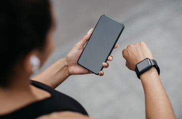 Device synchronization for sport. African american girl in sport bra and wireless headphones, looks at fitness tracker and smartphone in hands, free space