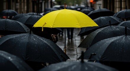 A bright yellow umbrella stands out amongst a sea of dark umbrellas on a rainy city street.