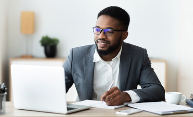 Obraz premium Handsome black entrepreneur using laptop and taking notes while working in modern white office, panorama