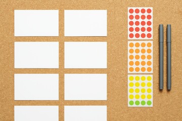 White blank cards, colorful round stickers in red, orange, yellow, green, and two pens arranged neatly on corkboard background