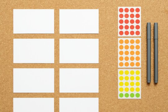 White blank cards, colorful round stickers in red, orange, yellow, green, and two pens arranged neatly on corkboard background