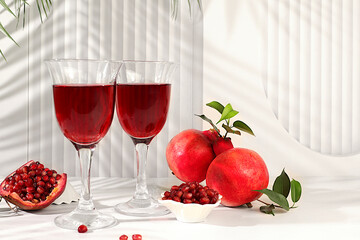 Glasses of red pomegranate wine and grains with ripe pomegranates on a trendy table with palm shades, concept festive party, advertising for a bar and cafe. Source of vitamins for a healthy lifestyle,