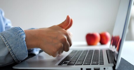 Woman wearing a denim jacket sits at a desk, typing on a laptop keyboard and giving a thumbs up gesture, with three apples visible in the background - Powered by Adobe