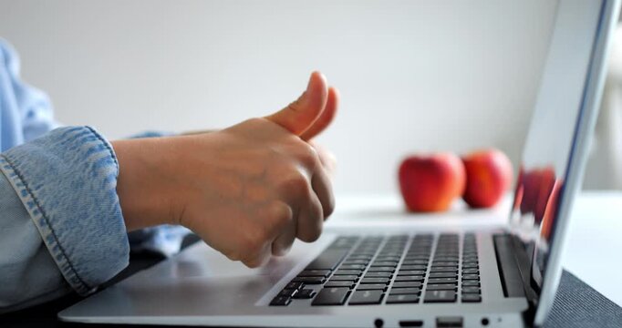 Woman wearing a denim jacket sits at a desk, typing on a laptop keyboard and giving a thumbs up gesture, with three apples visible in the background