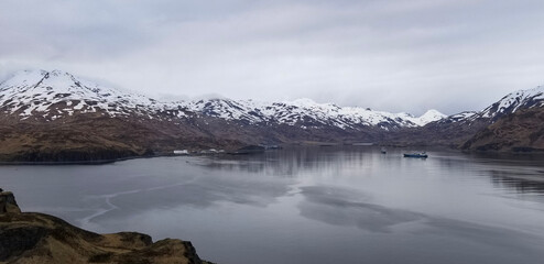 Dramatic winter landscape of the Unalaska in Alaska, showing snowy mountains, cold waters, and fishing vessels. A remote region known for its harsh climate and industrial fishing industry.