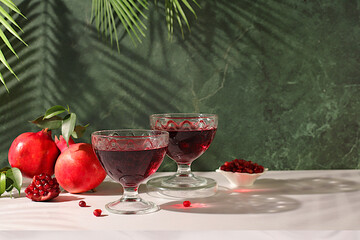 Glasses of red pomegranate wine and grains with ripe pomegranates on a trendy table with palm shades, concept festive party, advertising for a bar and cafe. Source of vitamins for a healthy lifestyle