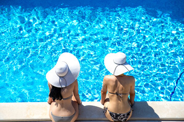 Top view of two young slim women siting, talking and resting on the edge of swimming pool in bathing suits and white straw sun hats during vacations. Travel and holiday concept, with copy space