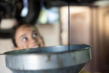 A young mechanic happily draining oil under a vehicle, showcasing automotive repair skills and confidence in her abilities. Selective focus on oil