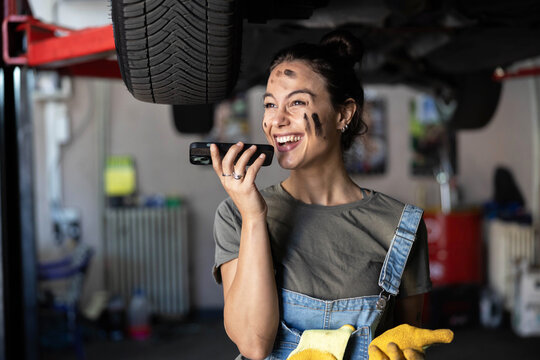 A laughing mechanic enjoys a phone conversation while working on a car in her tools-filled garage, radiating positivity.