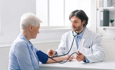 Fototapeta premium Young doctor measuring blood pressure of senior woman during visit at clinic