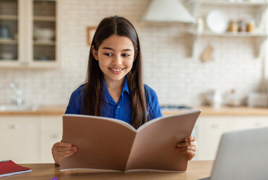 Homeschooling Education. Preteen Arab Girl Reading Text Book Doing School Homework At Home Interior. Middle Eastern Kid Schooler Studying Near Laptop While Sitting At Desk Indoor