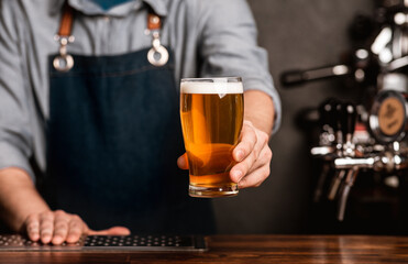 Fresh keg beer. Bartender in apron holds out a glass of light ale in interior of bar, cropped